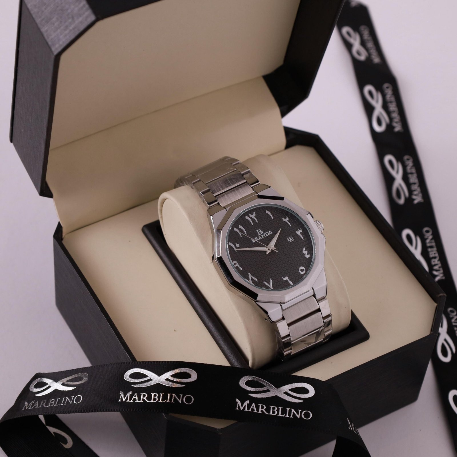 ساعة يد رجالية بسوار معدني فضي وقرص أسود بأرقام عربية شرقية، معروضة في علبة هدايا فاخرة بشريط ماربلينو (Marblino).