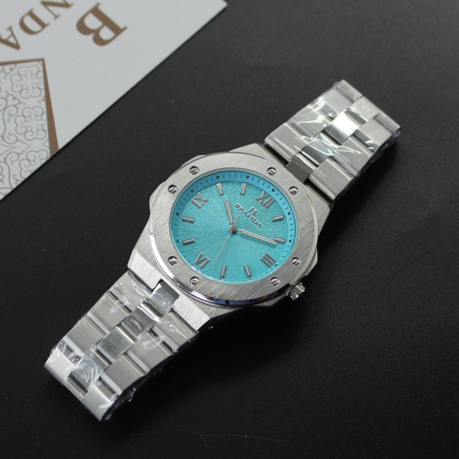 "ساعة يد (رجالية/للجنسين) بتصميم مثمن الأضلاع رياضية-فاخرة، بحافظة وسوار من الفولاذ المقاوم للصدأ فضي اللون، ومينا باللون الأزرق الفيروزي (Tiffany Blue) بنمط مزخرف، وعلامة تجارية BRAIDA."